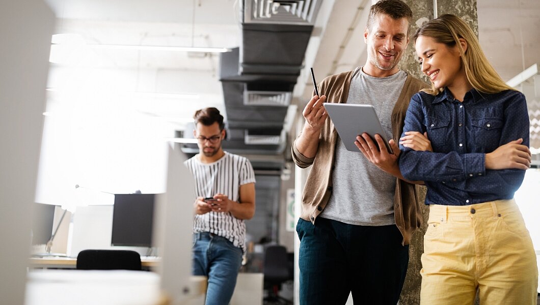 Zwei Personen stehen in einem Büro, schauen auf ein Tablet und besprechen etwas. Eine andere Person im Hintergrund sitzt und schaut auf ein Telefon.