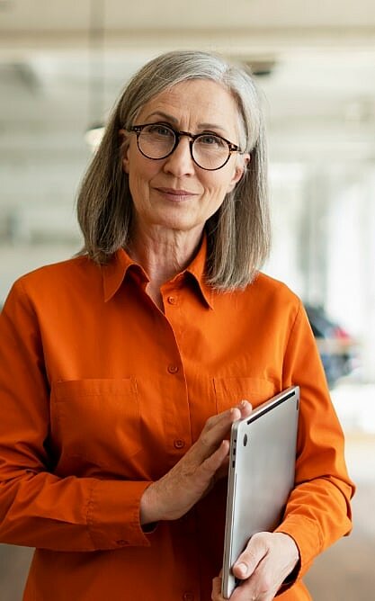Eine ältere Frau mit grauem Haar und Brille, die ein orangefarbenes Hemd trägt, hält ein Tablet und steht in einem Innenbereich.