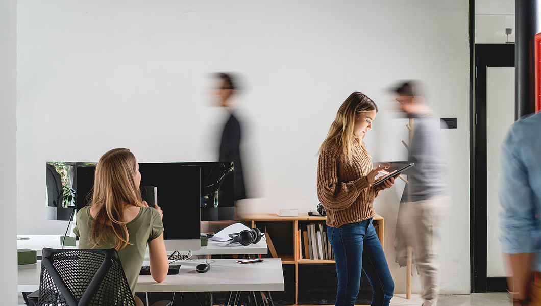 A woman is sitting at a desk in an office looking at a computer screen. Another woman stands next to it and reads something. Two blurred people are walking in the background.